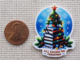 Small Christmas tree sitting on top of a stack of colorful books with a string of lights in front of a winter night circle with the words "All booked for Christmas" minder with a penny for scale.