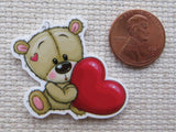 Minder of a bear holding a red heart next to a penny for size comparison on a textured surface.