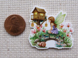 Minder of a fairy sitting on a flower with a small house in the background, next to a penny for scale.