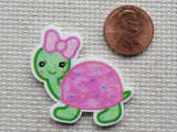 Cartoon turtle minder with pink shell and bow next to a penny on a textured surface
