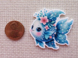 Decorative minder of a blue and white gold fish with floral elements next to a penny on a textured surface.