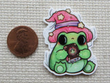 Cartoon frog with pink hat holding a book, next to a penny for scale, on a textured surface.