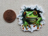 Minder of a green frog popping out from a hole next to a penny on a textured surface