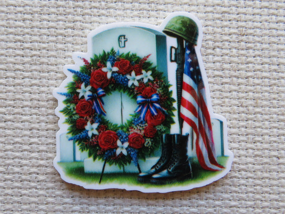 First view of a Simple military grave stone minder with a patriotic wreath hanging in front of it. A flag is next to it with military boots at the bottom and a helmet on the top of the flagpole.