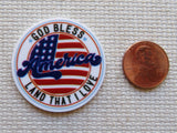 Round minder with 'God Bless America Land That I Love' text and an American flag design, next to a penny for scale.
