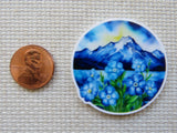 Minder with blue flowers and mountain design next to a penny on a textured surface