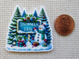 Second view of A cute snowman in front of a "Snowman Lane" sign in front of a snow covered forest minder.