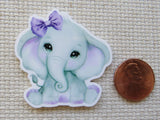 Minder of a cute elephant with a purple bow next to a penny on a textured surface
