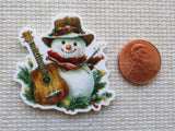 Minder of a snowman with a guitar next to a penny on a textured surface