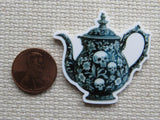 Minder of a teapot with skull design next to a penny on a textured surface