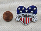 Heart-shaped 'God Bless America' minder with American flag design next to a penny on a textured surface.