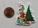 Minder of a green character with a red hat next to a Christmas tree and presents, compared in size to a penny on a textured surface.