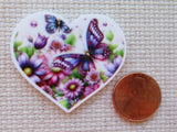 Heart-shaped minder with butterflies and flowers next to a penny on a textured surface