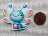 Cartoon dragonfly minder with colorful design on a textured surface next to a penny for scale.