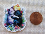Minder of three cats hugging with flowers, next to a penny for scale on a textured surface.