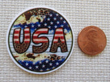 Round minder with 'USA' text and American flag design next to a penny on a textured surface