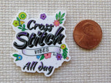 Minder with 'Cross Stitch Vibes All Day' text and flowers next to a penny for size comparison on a textured surface.