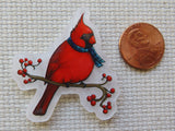 A minder featuring a red cardinal on a branch with red berries and a blue plaid scarf, shown next to a copper penny for scale on a textured white fabric background.