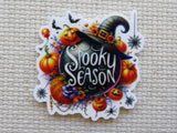 A scalloped Halloween-themed minder featuring the words 'Spooky Season' in white script on a dark oval, surrounded by orange pumpkins, a witch hat, bats, spiders, and cobwebs, shown on a beige woven fabric background.