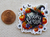 A scalloped white minder featuring a black witch hat, pumpkins, spiders, and the words 'Spooky Season' in white script, shown next to a U.S penny.