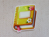 A minder shaped like a cute yellow and orange book with pink and white flowers, shown on a beige textured fabric background.