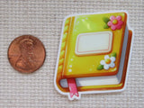 A minder shaped like a small illustrated book or notebook with a yellow cover, orange border, pink ribbon bookmark, and flower details.