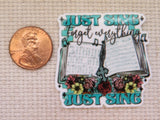 A minder featuring the text 'Just Sing Forget Everything Just Sing' with an open book, musical notes, and floral accents, shown next to a copper penny on a beige textured fabric background.
