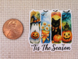 A Halloween-themed minder with four vertical panels showing a ghost with a pumpkin, daisies with autumn leaves, a black cat with a bow tie, and a haunted house, with the text "Tis The Season" below. A copper penny is placed to the left for scale on a beige textured fabric background.