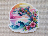 A minder featuring a colorful tattoo-style dolphin surrounded by pink hibiscus flowers and blue ocean waves, set against a pink sunset background, displayed on a beige woven fabric surface.