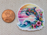 A minder featuring a colorful dolphin with flowers, pink hibiscus flowers, and blue waves against a sunset background, shown next to a US penny for scale on a beige woven fabric background.