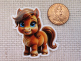 A minder featuring a cute cartoon pony with brown hair and blue eyes, shown next to a US penny for scale on a textured beige fabric background.