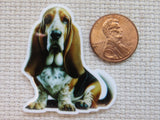 A minder of a Basset Hound dog shown next to a US penny for scale, placed on a textured beige fabric background.