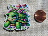 A minder featuring a cute green turtle surrounded by pink flowers and mushrooms, shown next to a US penny on a textured beige fabric background.